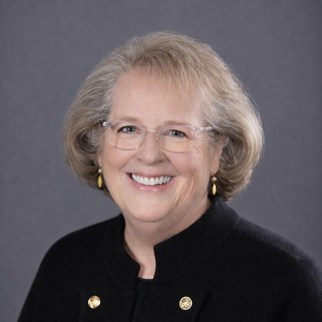 Professional headshot of Carol Gieske smiling with short, light brown and gray hair, wearing glasses and a black jacket with gold buttons, set against a neutral gray background.