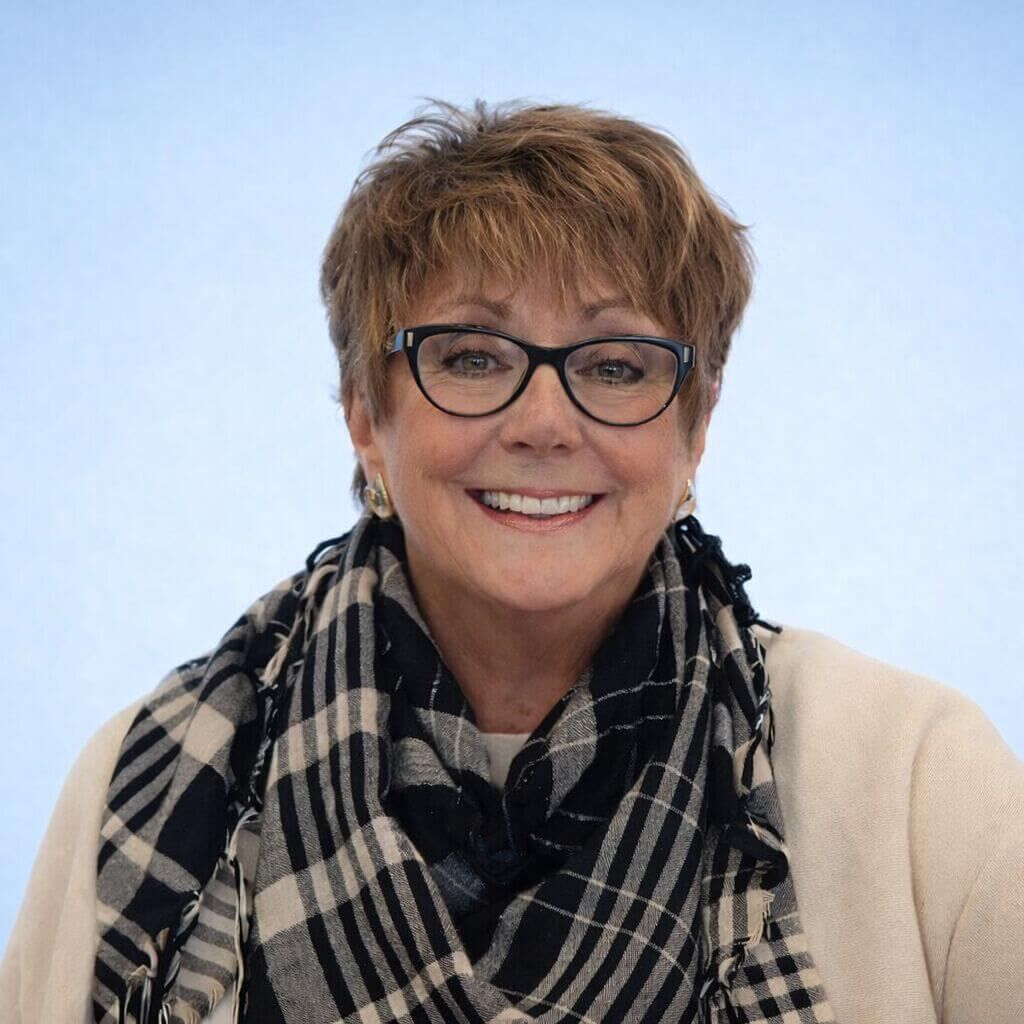 Professional headshot of Linda Siete smiling with short brown hair and glasses, wearing a cream top and plaid scarf, set against a soft light blue background.