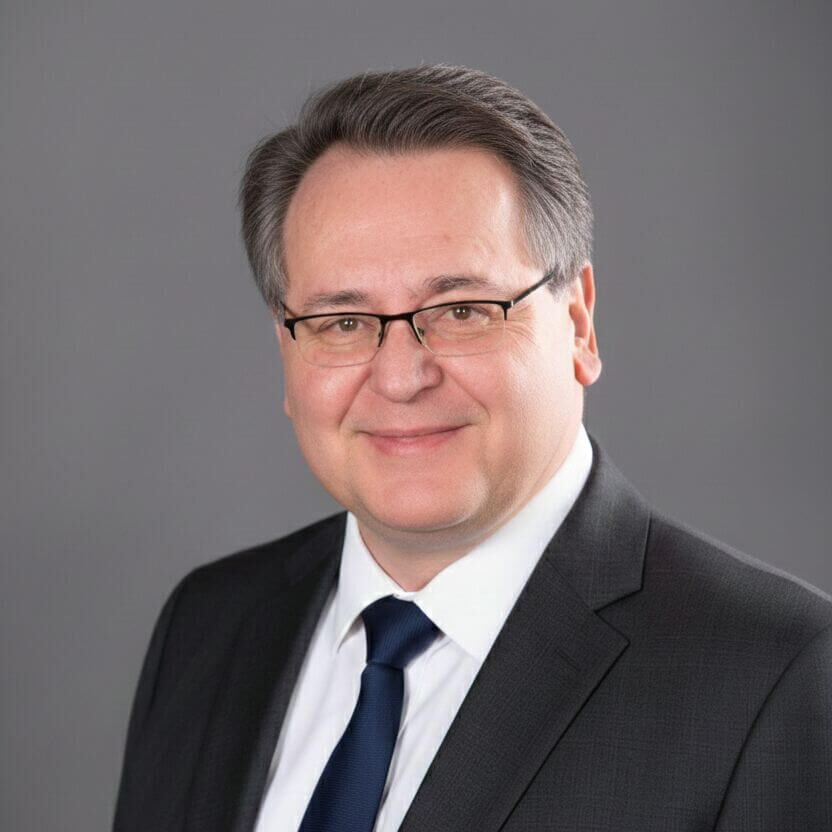 Professional headshot of Tony Lucenko wearing glasses, a dark suit, white shirt, and tie, smiling slightly against a neutral background.